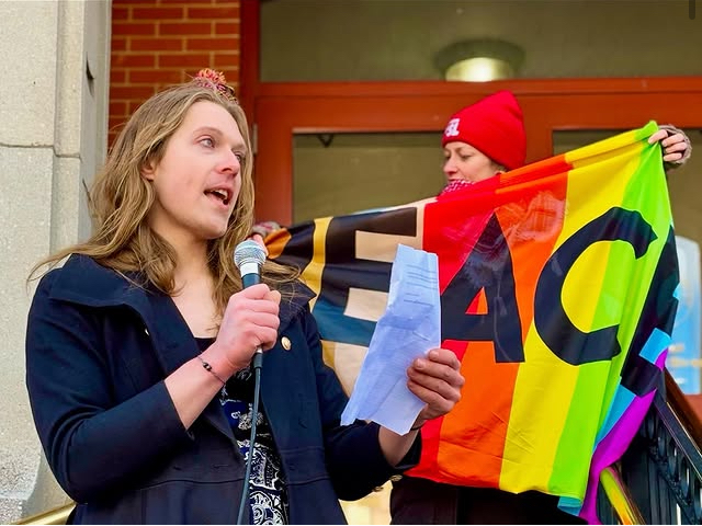 Image of speaker at a rally, with rainbow peace flag behind. 
