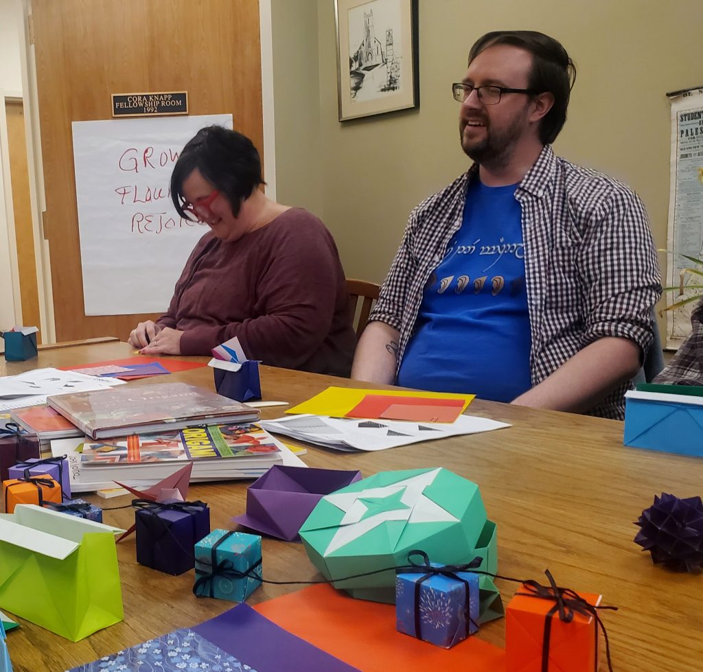 Two individuals sit at a table. A woman with a purple shirt folds origami. A man with a blue shirt smiles. Origami boxes and other papercrafts are spread out on the table top.