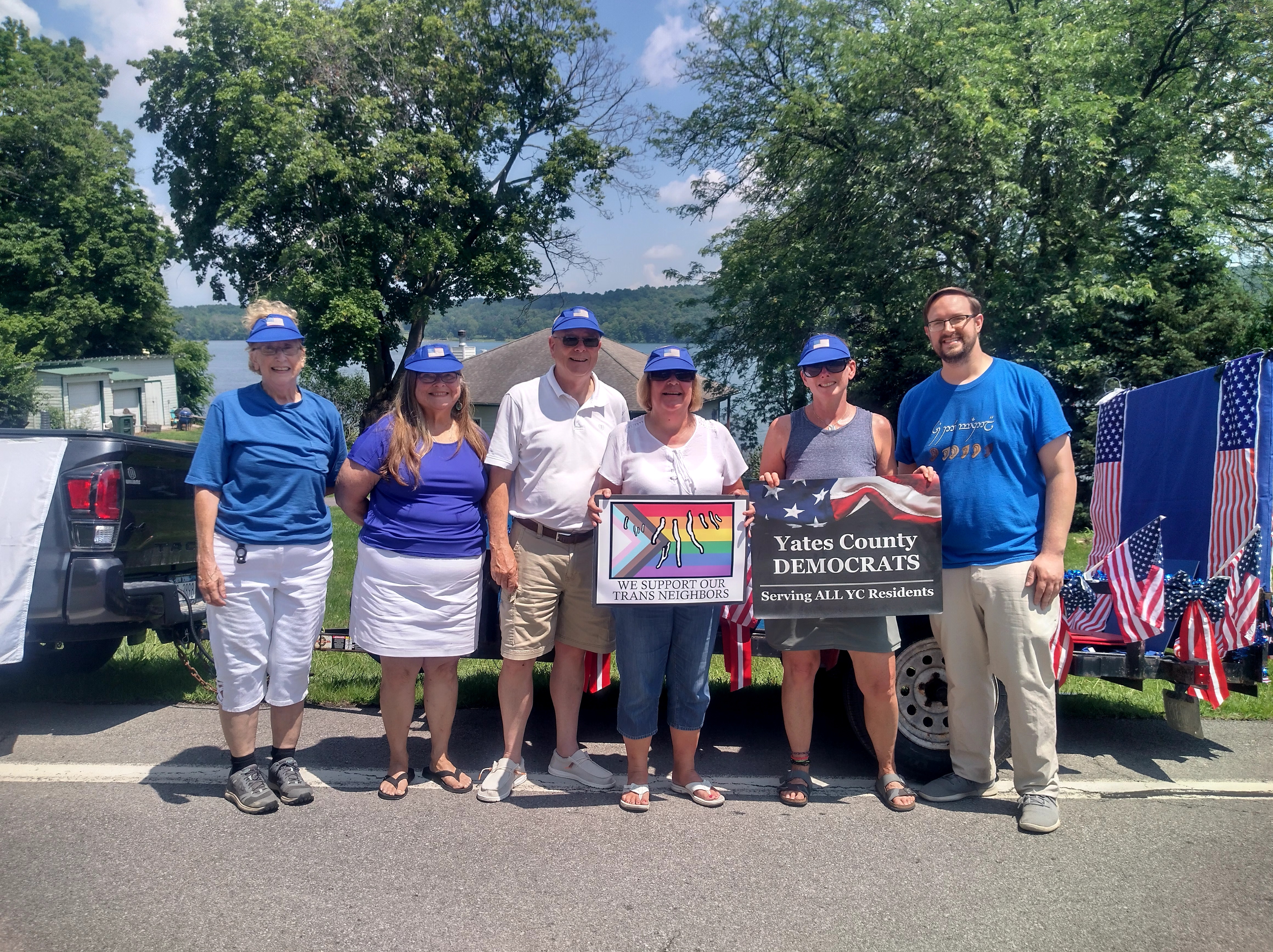 A group of people poses for a photo. They are holding two signs. They read, "We Support our Trans Neighbors," and "Yates County Democrats, Serving ALL YC Residents."