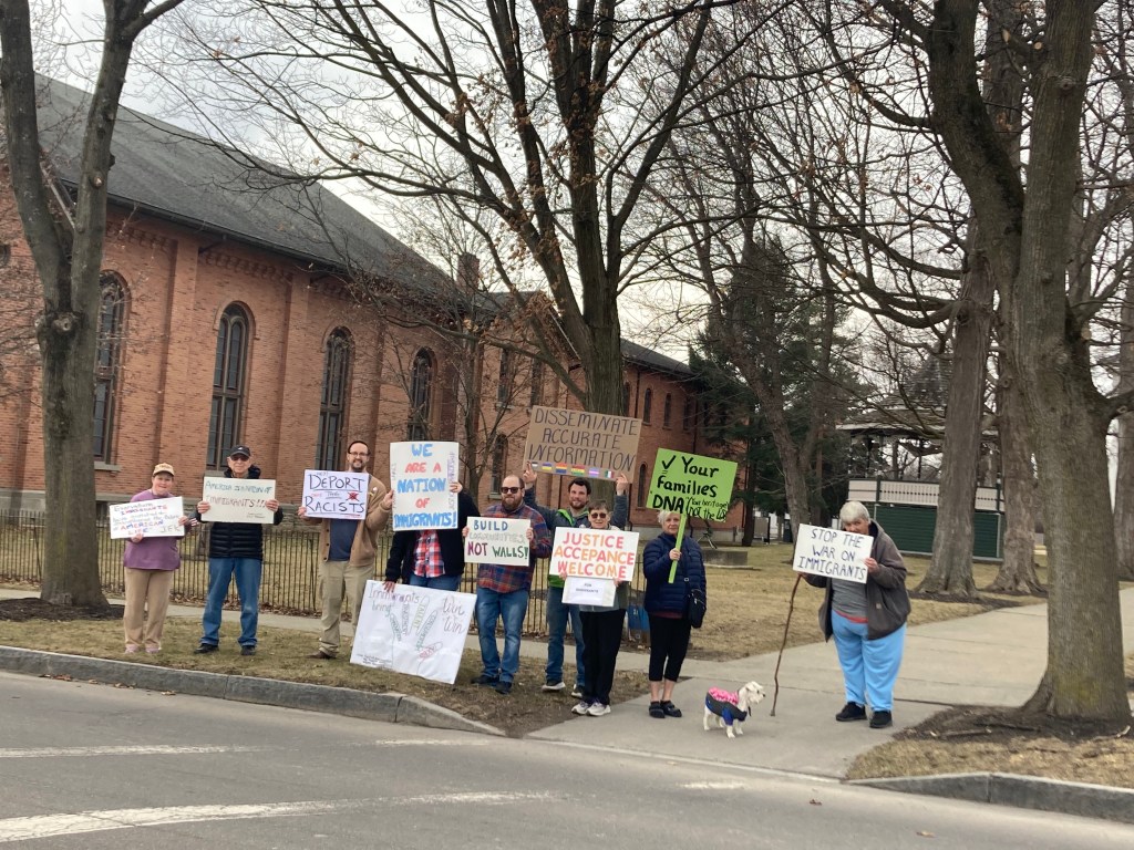 A photo of individuals standing on the side of the road holding signs in a rally.