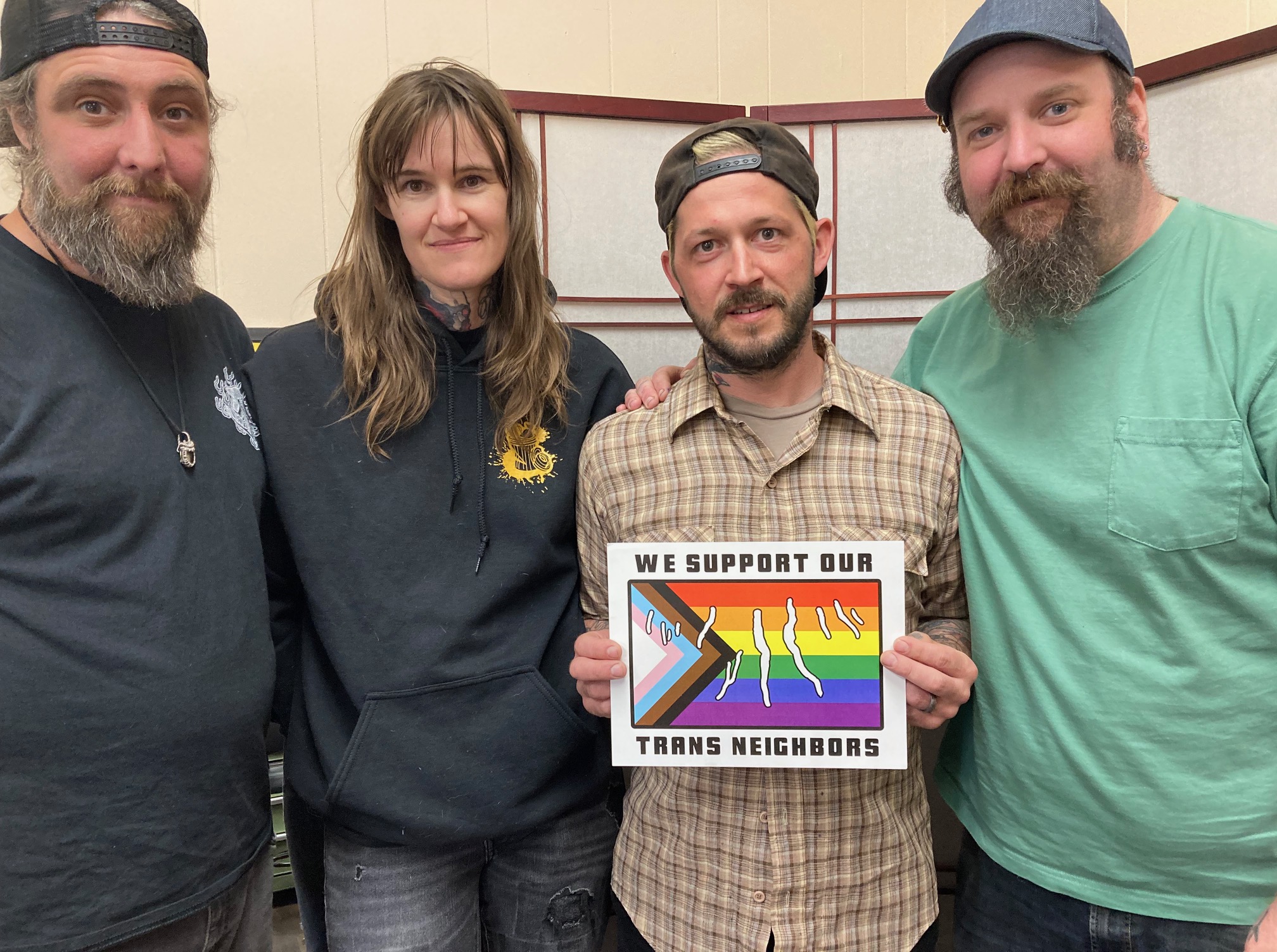 A group of people stands. One person holds a sign which reads, "We Support Our Trans Neighbors." It has an outline of the Finger Lakes on top of an image of the progress pride flag.