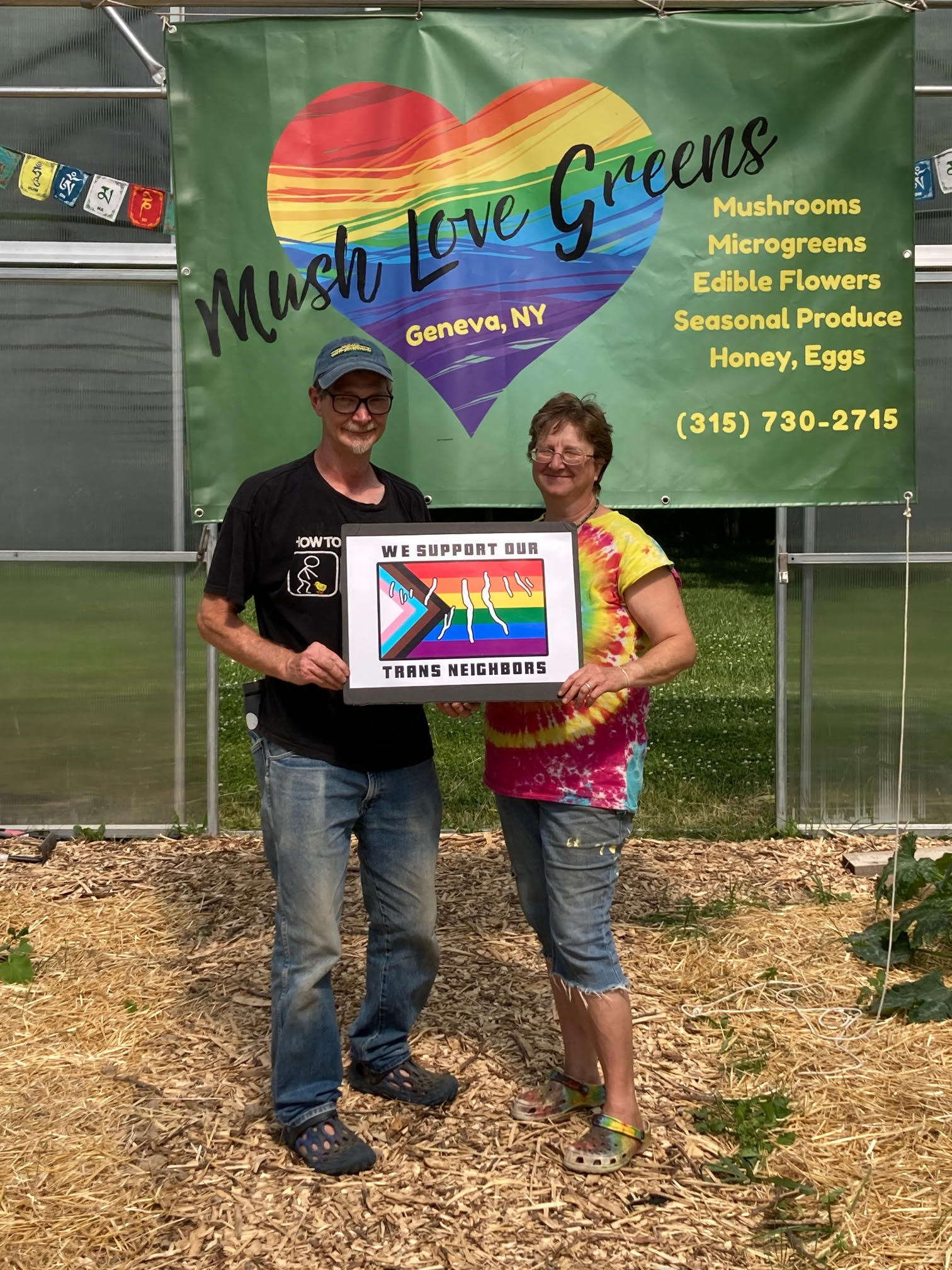 Two people holding a sign which reads, "We Support our Trans Neighbors." They are standing in front of a banner which reads, "Mush Love Greens, Geneva, NY, Mushrooms, Microgreens, Edible Flowers, Seasonal Produce, Honey, Eggs, (315) 730-2715."